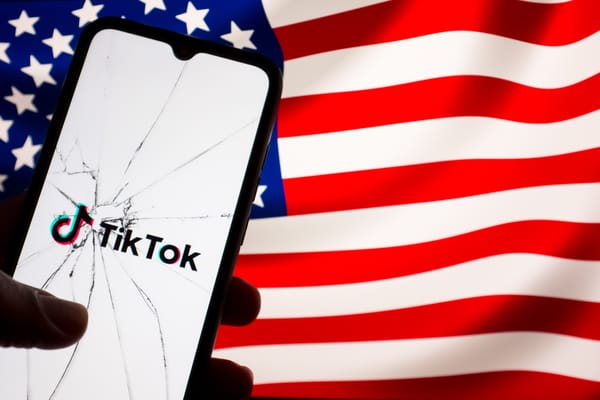 The TikTok logo displayed on a phone seen through broken glass, with a US flag appearing on a screen in the background